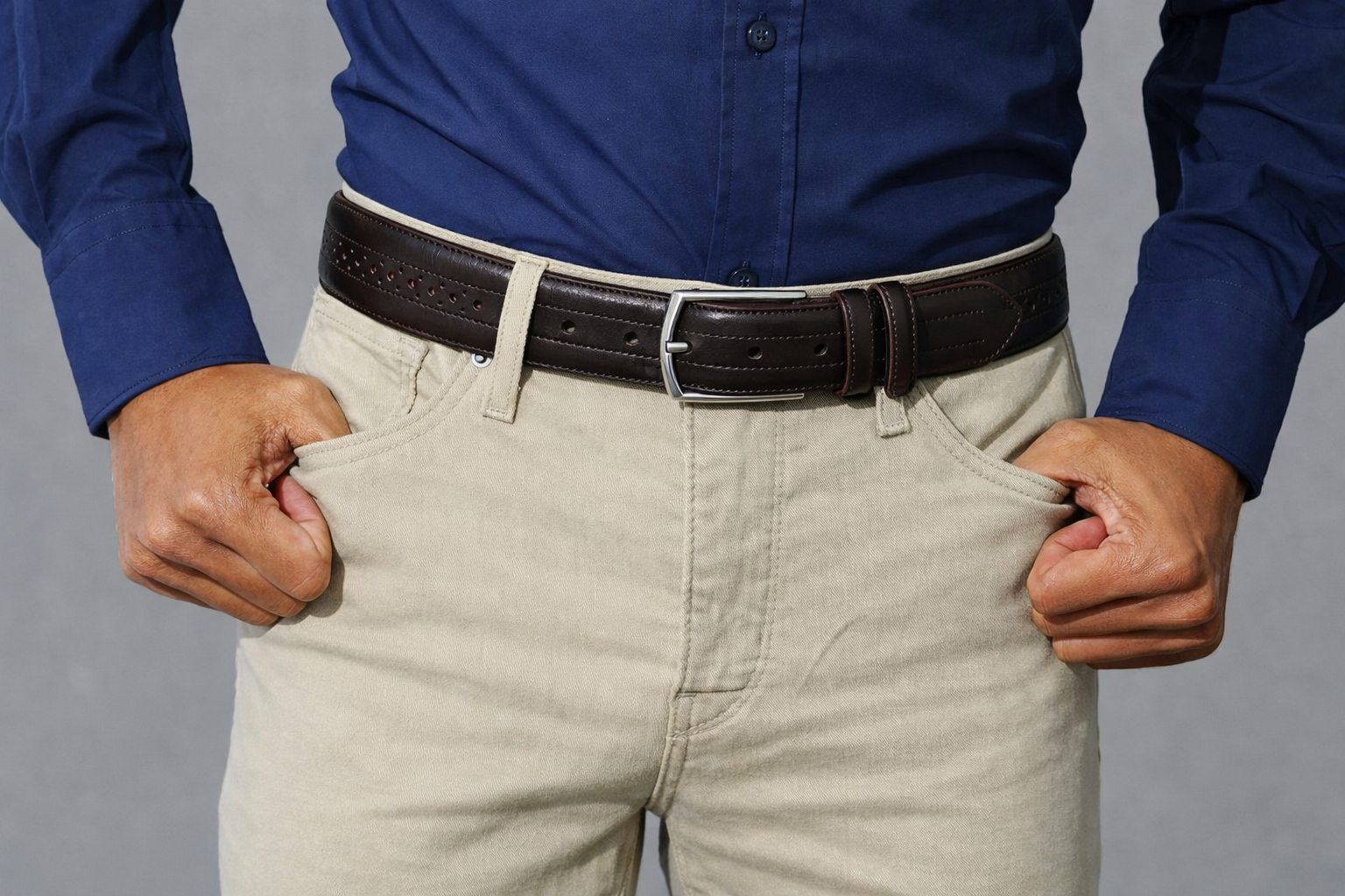 Close up of a man wearing a 35mm dark brown full grain calfskin brogue belt with stone cotton chinos and a navy blue long sleeve button up shirt.